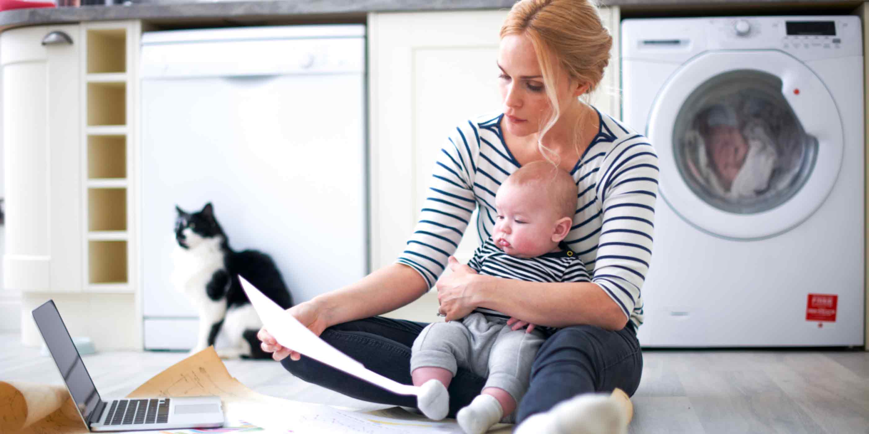 woman sitting next to her laptop and cat in laundry room holding baby and reading a page of something tha tlooks interesting