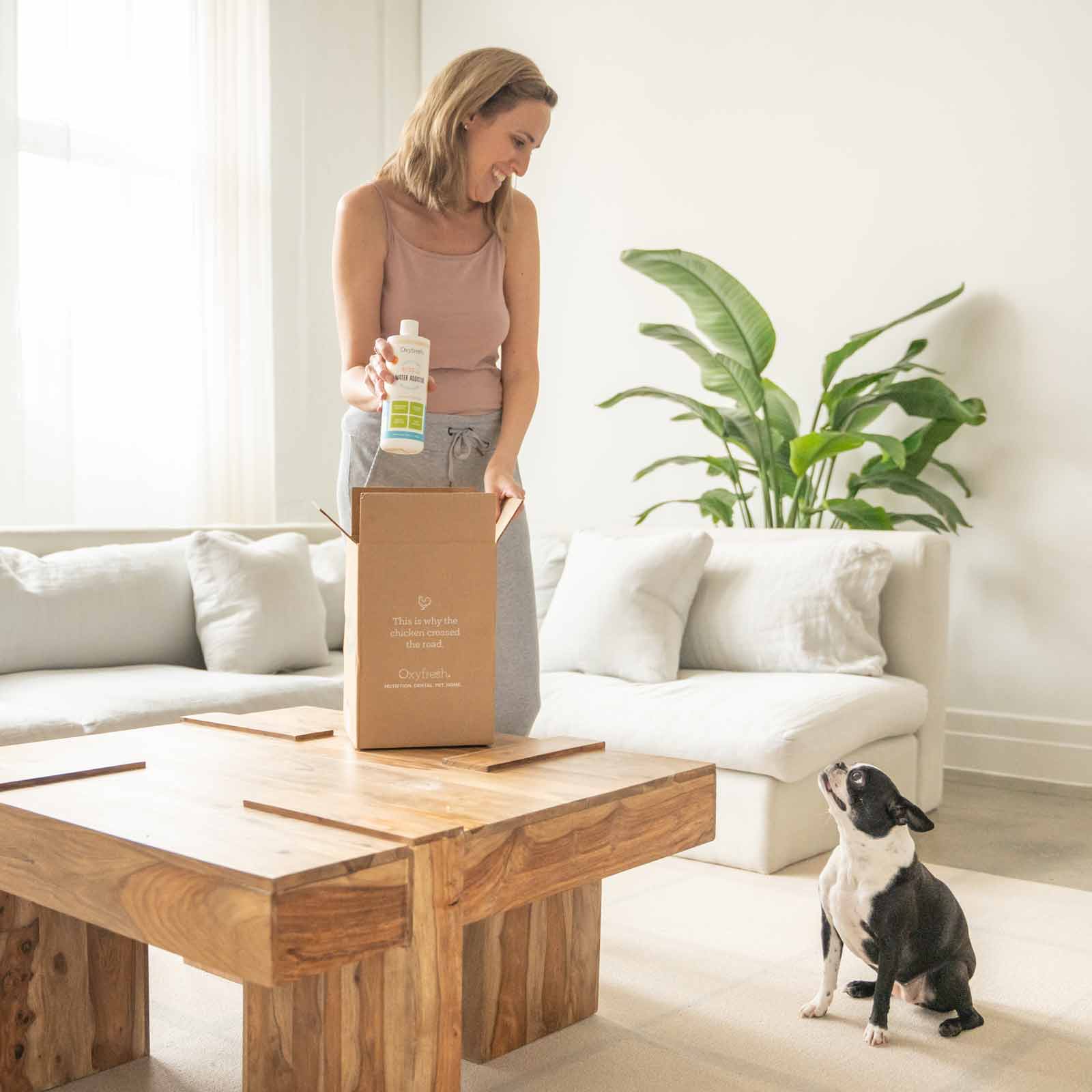 woman smiling down at her pup as she pulls a bottle of oxyfresh pet water additive for fresh dog breath out of the box