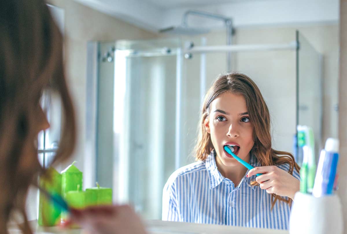 oxyfresh-dental-blog-titled-Why-Is-My-Toothpaste-Burning-My-Mouth_What-You-need-to-know_Woman-brushing-her-teeth-looking-questioningly-into-the-mirror