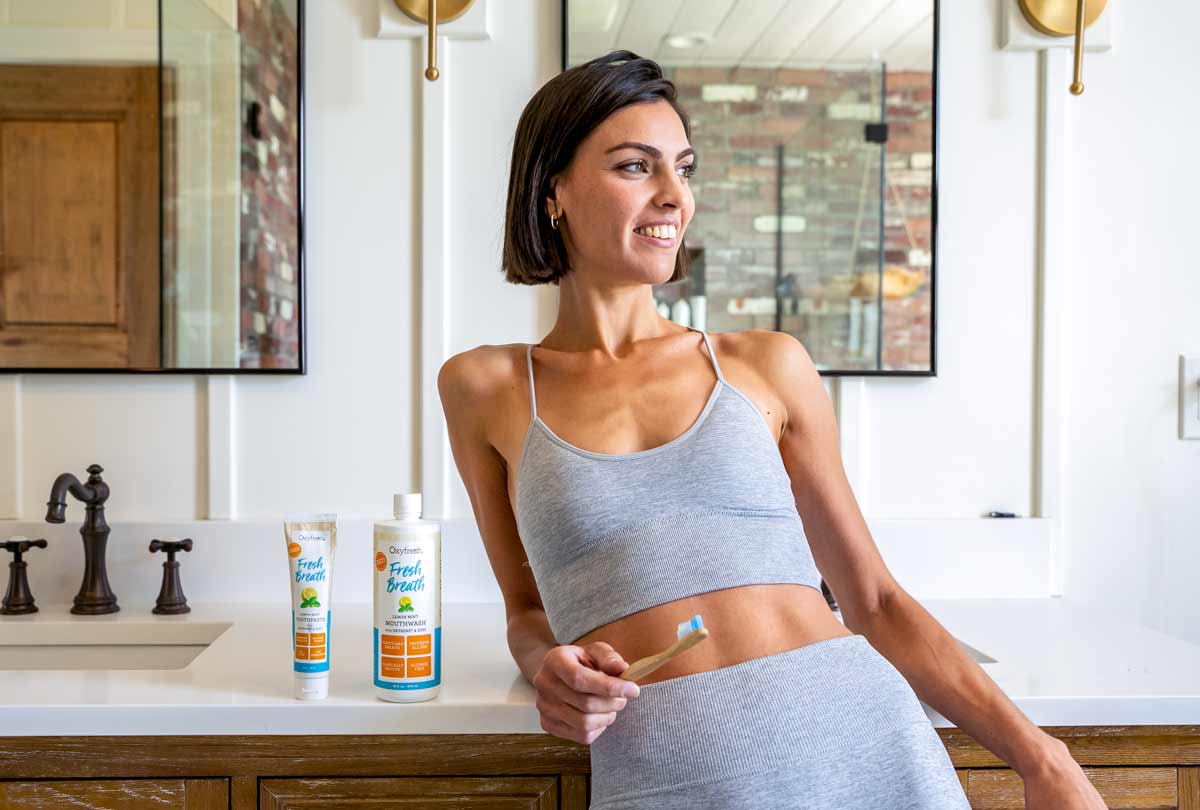 woman-smiling-and-casually-leaning-on-bathroom-counter-with-oxyfresh-lemon-mint-mouthwash-and-toothpaste-next-to-her