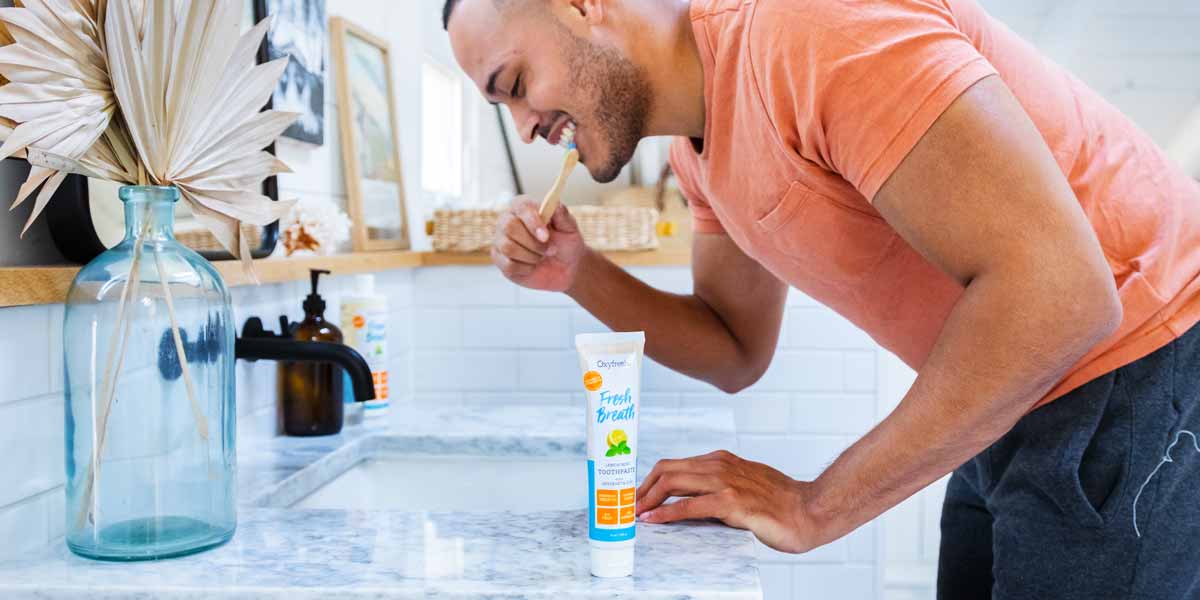 Man-bending-over-sink-while-brushing-his-teeth-in-modern-bright-white-bathroom-with-oxyfresh-lemon-mint-toothpaste-on-the-counter-beside-him