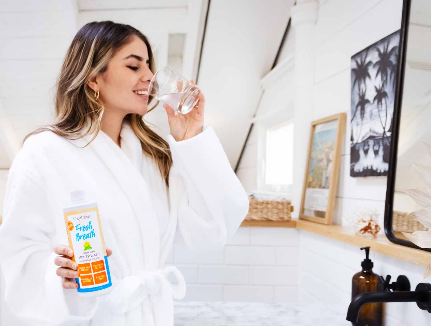 Woman-in-a-white-bathrobe-with-a_bright-bathroom-raising-mouthrinse-glass-to-her-lips-as-she-smiles-and-holds-a-bottle-of-lemon-mint-mouthwash-in-her-other-hand