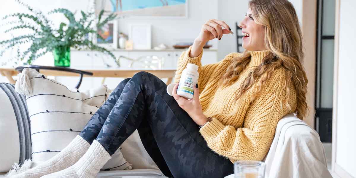 Woman-sitting-on-couch-looking-up-smiling-and-taking-a-vitality-capsule-with-workout-clothes