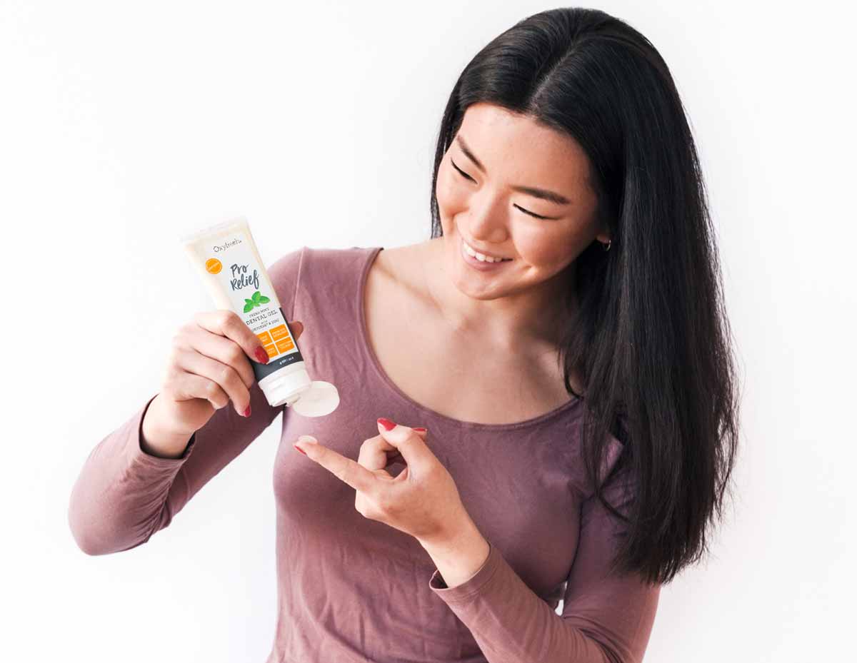Woman-with-a-dab-of-oxyfresh-pro-relief-gel-smiling-and-raising-her-hand-to-her-mouth-to-apply-the-dental-gel-to-her-gums