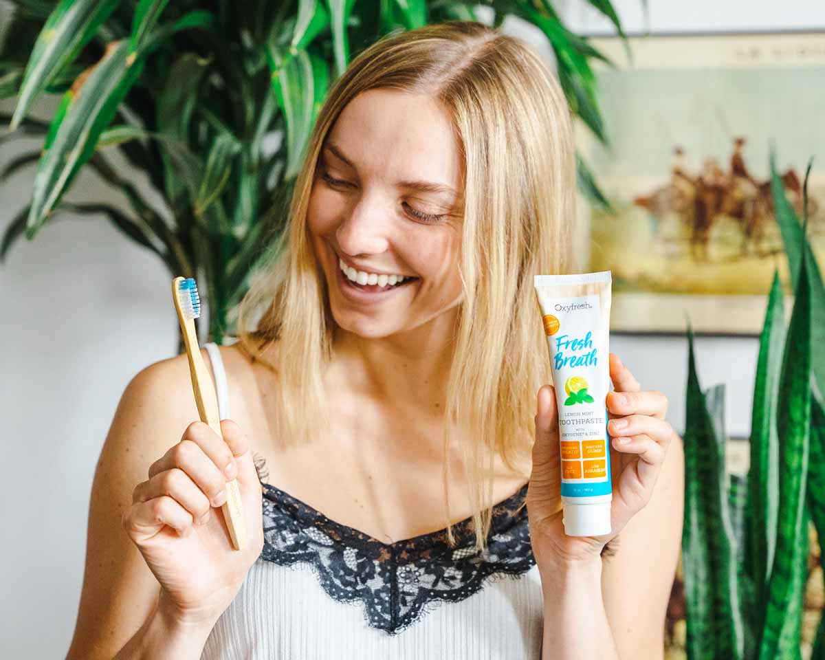 Woman-with-a-dazzling-white-smile-holding-oxyfresh-lemon-mint-toothpaste-and-a-dense-bristled-bamboo-toothbrush-in-cute-boho-apartment-with-deep-green-plants-in-the-background