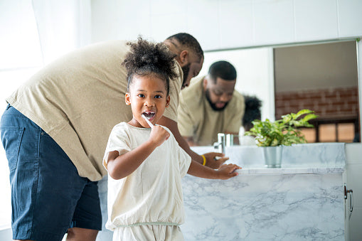 little girl looking at the camera while brushing her teeth with dad