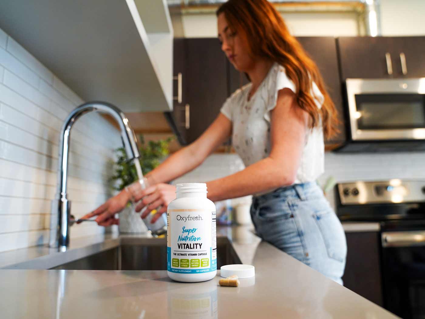 woman-pouring-water-into-a-glass-preparing-to-take-an-oxyfresh-super-nutrition-vitality-capsule-which-is-laying-on-the-counter-in-the-foreground